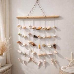 May include: A wall hanging featuring a wooden dowel from which multiple strands of twine are suspended. Various seashells in shades of white, brown, and tan are attached to the twine, creating a coastal-themed decorative piece.