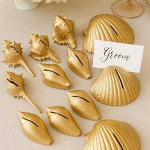 May include: Gold-painted seashell place card holders, perfect for a beach-themed wedding or event. The shells are in various shapes and sizes, with a slot to hold a card. One card reads "Groom".