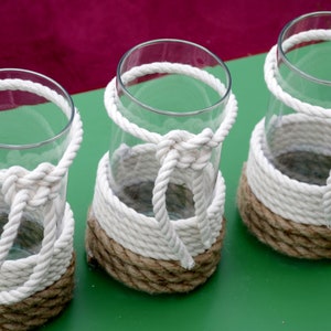 May include: Three clear glass vases with a nautical design. Each vase is wrapped with thick, white rope and a natural-colored rope. The rope is knotted around the vase, creating a decorative and functional element.