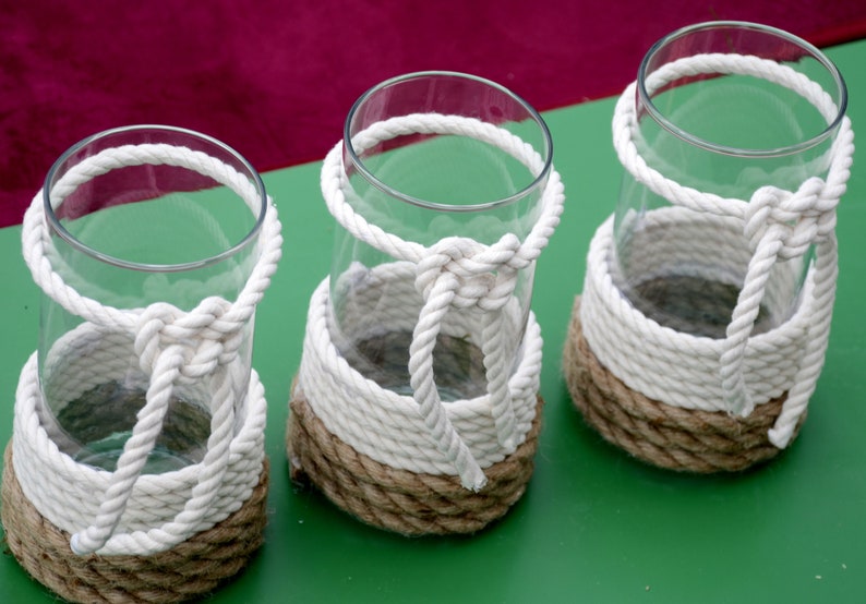 May include: Three clear glass vases with a nautical design. Each vase is wrapped with thick, white rope and a natural-colored rope. The rope is knotted around the vase, creating a decorative and functional element.