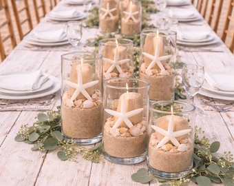 May include: A beach-themed table setting with white plates, glasses, and candles in glass cylinders. Each candle is adorned with a starfish and set in sand and seashells. Greenery decorates the table, which is made of white-painted wood.
