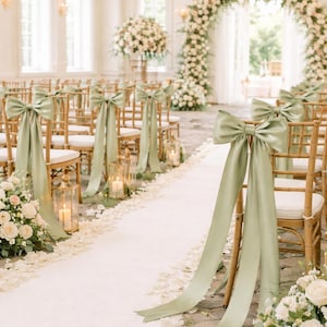 May include: Elegant wedding ceremony setup with rows of gold chairs adorned with sage green bows and ribbons. A white aisle runner is flanked by floral arrangements and lanterns, leading to a floral arch.