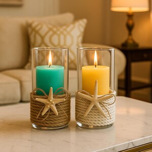 May include: Two cylindrical glass candle holders, each containing a lit pillar candle. One candle is teal with a brown rope wrap and a beige starfish. The other candle is yellow with a white rope wrap and a beige starfish attached to the front.