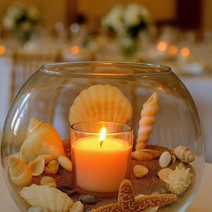 May include: A glass bowl filled with sand, seashells, and a lit orange candle. The candle is in a clear glass holder. A starfish is visible. The background is blurred, suggesting a wedding or event setting.