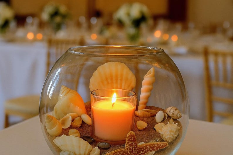 May include: A glass bowl filled with sand, seashells, and a lit orange candle. The candle is in a clear glass holder. A starfish is visible. The background is blurred, suggesting a wedding or event setting.