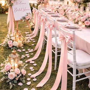 May include: A white welcome sign with the text Welcome to the Bridal Shower, decorated with pink roses. White chairs feature long pink ribbons tied in bows. Gold lanterns with candles and pink floral arrangements are placed on the grass.
