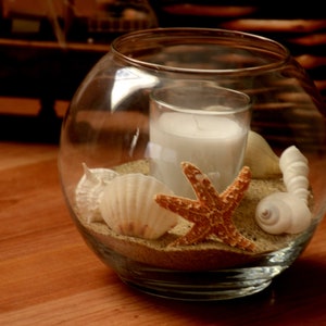 May include: A clear glass bowl containing a white candle, sand, seashells, and a starfish. The candle is in a clear glass holder. The seashells are white and beige, and the starfish is orange. The bowl rests on a wooden surface.