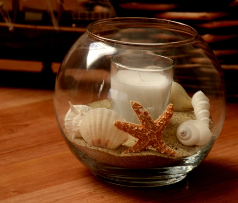 May include: A clear glass bowl containing a white candle, sand, seashells, and a starfish. The candle is in a clear glass holder. The seashells are white and beige, and the starfish is orange. The bowl rests on a wooden surface.