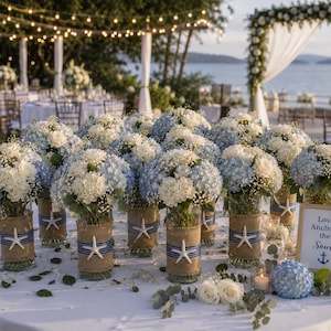 May include: A collection of glass vases filled with white and light blue hydrangea bouquets, wrapped in burlap and navy blue ribbon with white starfish accents. A framed sign reads Love Anchors the Soul with an anchor icon on a white table.
