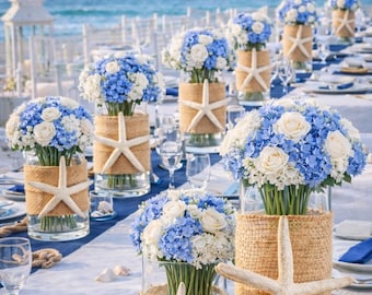 May include: Beach-themed table setting with blue and white floral arrangements in glass vases wrapped with burlap and adorned with starfish. The table is set with blue and white linens, seashells, and rope, with the ocean in the background.