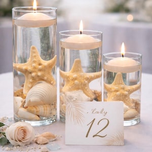May include: Three glass cylinders with floating candles, starfish, and seashells. A table number card with the number 12 is in front of the cylinders. The scene is set on a white tablecloth, with a soft focus background.