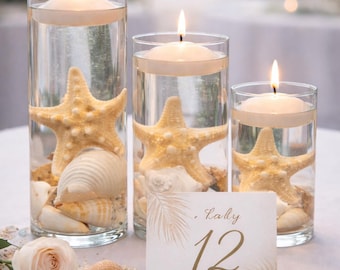 May include: Three glass cylinders with floating candles, starfish, and seashells. A table number card with the number 12 is in front of the cylinders. The scene is set on a white tablecloth, with a soft focus background.