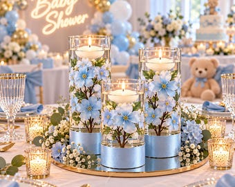 May include: Three cylindrical glass vases with light blue floral decals and metallic blue bases, each containing a floating white candle. The vases are arranged on a circular gold mirror tray surrounded by small white flowers and greenery.