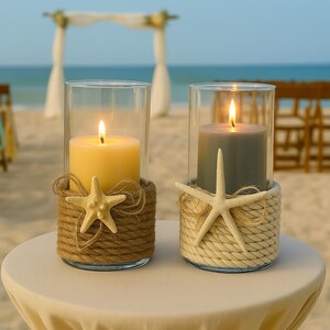 May include: Two cylindrical glass candle holders wrapped in rope. The left holder features brown rope and a yellow candle, while the right holder has cream rope and a gray candle. Each is decorated with a single beige starfish and twine.