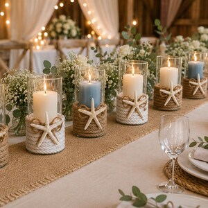 May include: A series of cylindrical glass candle holders wrapped in natural jute rope and white cord, each featuring a white starfish accent. The holders contain lit pillar candles in shades of light blue and cream, arranged on a burlap table runner.