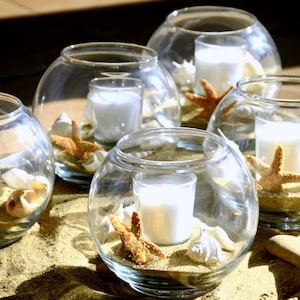 May include: Clear glass globe candle holders filled with sand, seashells, and starfish. Each globe holds a small white candle. The arrangement is set on a sandy surface, creating a beach-themed decorative display.