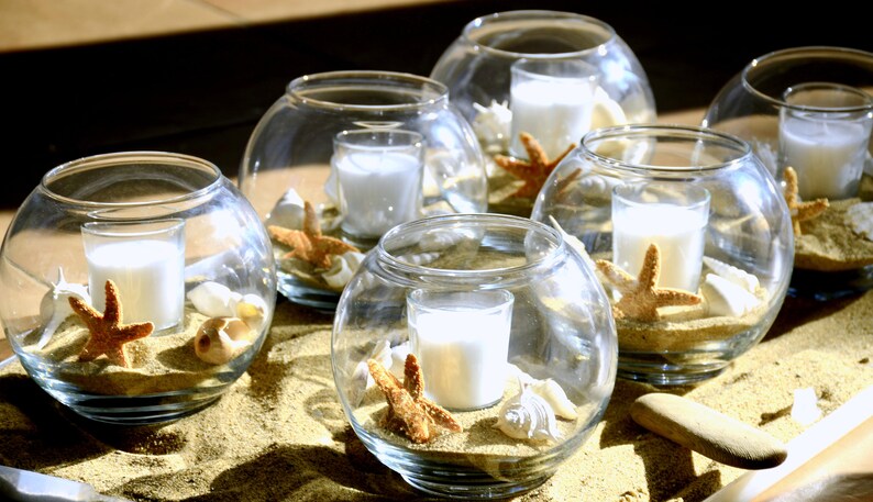May include: Clear glass globe candle holders filled with sand, seashells, and starfish. Each globe holds a small white candle. The arrangement is set on a sandy surface, creating a beach-themed decorative display.