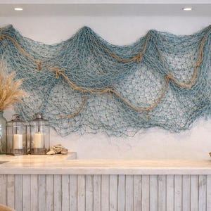 May include: Coastal-themed decor featuring a large blue fishing net hanging on a white wall. The display includes a glass vase with dried pampas grass, lanterns with candles, and a woven tray with candles. A wicker basket holds a notepad and pen.