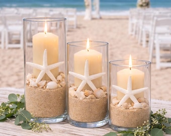 May include: Three glass cylinder candle holders with lit ivory candles, sand, seashells, and white starfish. The candles sit on a weathered wooden table with greenery, with a beach and wedding arch in the background.