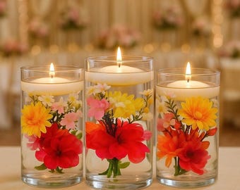 May include: Three clear cylindrical glass vases filled with water, each containing submerged colorful artificial flowers in shades of red, orange, yellow, and pink, topped with a single white floating candle with a lit flame.