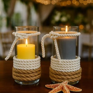 May include: Two glass candle holders with lit candles, one yellow and one gray, are decorated with rope. The base and top of the holders are wrapped in thick, brown rope, with white rope accents. A starfish rests on the table.