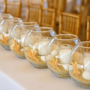 May include: Several clear glass bowls filled with sand, seashells, and starfish. Each bowl holds a white candle. The bowls are arranged on a white surface, with wooden chairs in the background. The theme is beach decor.