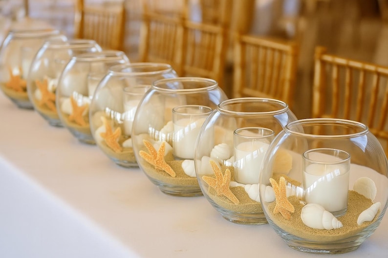 May include: Several clear glass bowls filled with sand, seashells, and starfish. Each bowl holds a white candle. The bowls are arranged on a white surface, with wooden chairs in the background. The theme is beach decor.