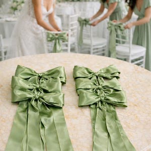 May include: Two sage green satin chair sashes, each featuring a large decorative bow with long, flowing tails. The fabric has a smooth, reflective finish, and the sashes are laid flat on a round table with a light-colored, marbled surface.