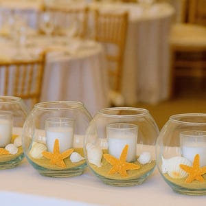May include: Glass bowl centrepieces with candles, sand, seashells, and orange starfish decorations. These beach-themed decorations are ideal for weddings or special events. The candles are white and cylindrical.