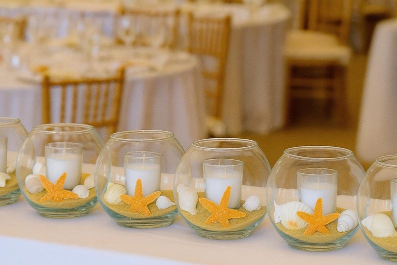 May include: Glass bowl centrepieces with candles, sand, seashells, and orange starfish decorations. These beach-themed decorations are ideal for weddings or special events. The candles are white and cylindrical.