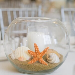 May include: A clear glass bowl containing sand, seashells, and an orange starfish. A white candle is inside a clear glass cylinder in the centre. The arrangement has a beach theme.