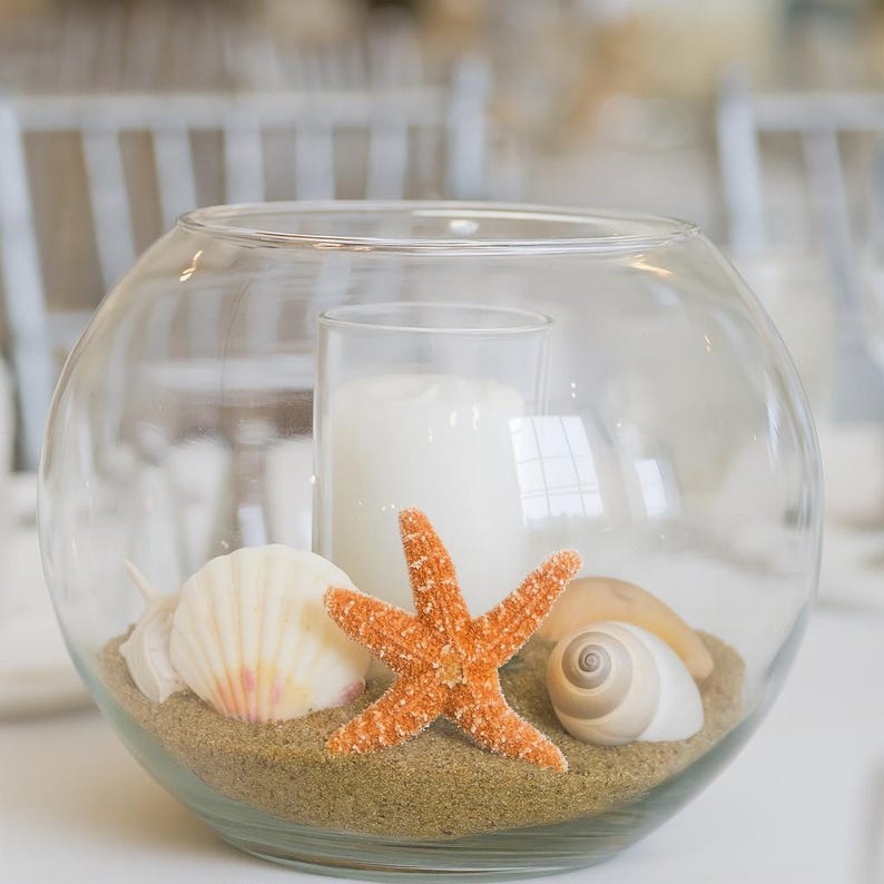 May include: A clear glass bowl containing sand, seashells, and an orange starfish. A white candle is inside a clear glass cylinder in the centre. The arrangement has a beach theme.