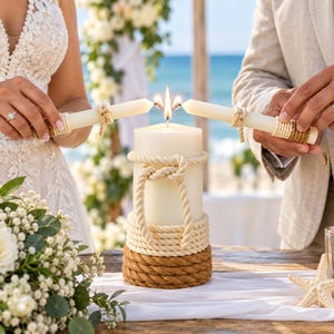 May include: A large white pillar candle wrapped in thick cream and brown nautical rope, featuring a decorative knot. Two smaller white taper candles are held by two individuals, with their flames meeting to light the central pillar candle.