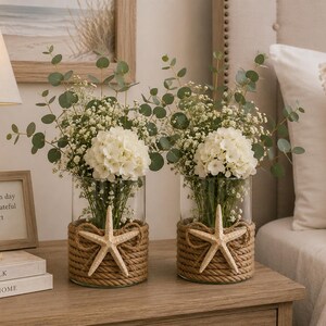 May include: Two clear glass cylindrical vases wrapped in thick natural jute rope, each featuring a single beige starfish accent. The vases are filled with white hydrangea blossoms, delicate baby's breath, and green eucalyptus stems.