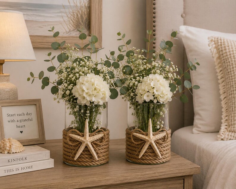 May include: Two clear glass cylindrical vases wrapped in thick natural jute rope, each featuring a single beige starfish accent. The vases are filled with white hydrangea blossoms, delicate baby's breath, and green eucalyptus stems.
