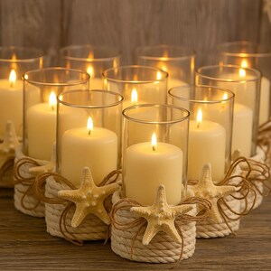 May include: A set of eight cylindrical glass candle holders, each containing a cream-colored pillar candle. The base of every holder is wrapped in thick, light-colored rope and decorated with a single beige starfish tied with twine.