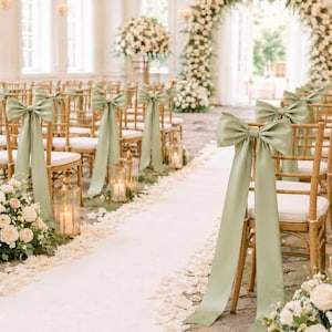 May include: A set of wooden chairs with white cushions, each adorned with a large sage green satin ribbon tied in a bow on the backrest. The chairs are arranged in rows along a white aisle runner decorated with scattered white flower petals.