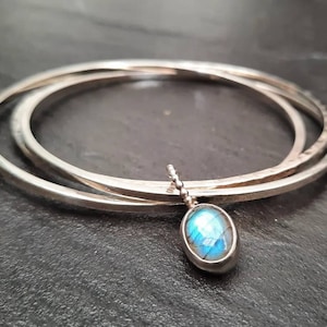 Könnte beinhalten: Ein silbernes Armreif-Armband mit drei Bändern, mit einem silbernen Anhänger mit einem blauen Labradorit-Edelstein.