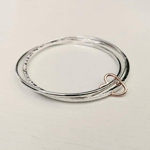 Könnte beinhalten: Drei silberne Armreifen mit einem roségoldenen Herz-Anhänger. Die Armreifen sind übereinander gestapelt und haben eine gehämmerte Textur.