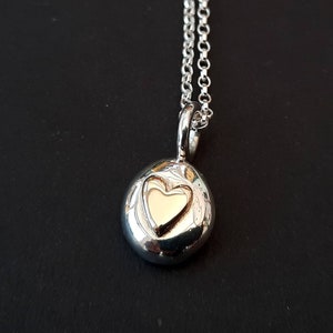 May include: A silver necklace with a round pendant featuring a small gold heart. The pendant has a textured surface.