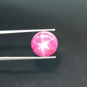 Peut inclure: Une gemme rose polie et ronde avec un motif en forme d'étoile. La gemme est maintenue par une pince à épiler argentée sur un fond sombre. L'effet d'étoile est créé par la lumière réfléchie par les inclusions dans la pierre.