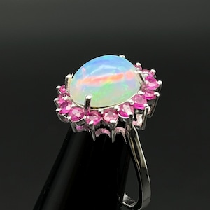 Peut inclure: Bague en argent ornée d'une opale ovale irisée, entourée d'un halo de petites pierres roses. Le bijou est présenté sur un fond sombre, mettant en valeur les couleurs de la pierre.