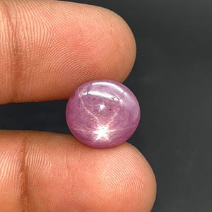 Puede incluir: Una gema redonda y pulida de color rosa con un efecto de estrella. La gema se sostiene entre dos dedos sobre un fondo oscuro. El efecto de estrella se crea por la reflexión de la luz en las inclusiones de la piedra.