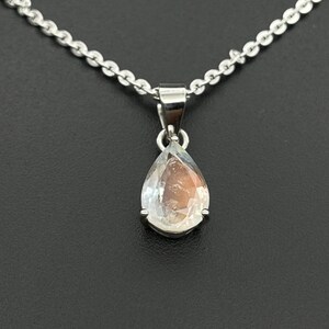May include: A silver necklace with a teardrop-shaped pendant. The pendant features a clear, iridescent gemstone. The chain is delicate and the pendant is centered against a dark background. The necklace is a piece of jewelry.