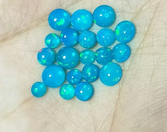 May include: A collection of 18 small, round, iridescent opal cabochons in shades of blue and green. The cabochons are arranged in a loose pile on a white surface.