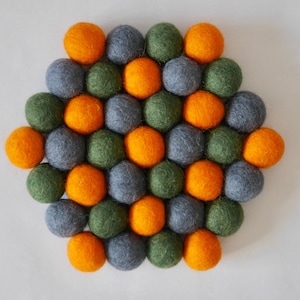 Puede incluir: Un posavasos redondo de fieltro hecho de bolas naranjas, verdes y grises. El posavasos está dispuesto en un patrón hexagonal.
