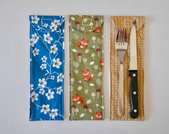 Peut inclure: Trois pochettes en tissu avec des motifs différents. La pochette de gauche a un fond bleu avec des fleurs blanches. La pochette du milieu a un fond vert avec des renards orange et des champignons. La pochette de droite a un fond jaune avec un motif blanc et contient une fourchette et un couteau.