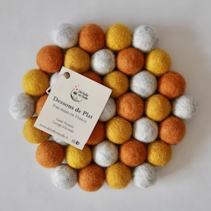 Op de afbeelding: Een ronde, vilt onderzetter gemaakt van witte, gele en oranje pompoenen. De onderzetter heeft een klein label met de tekst "Dessous de Plat" in het Frans, "Fait main en France", "Laine Feutrer", "Lavage à la main" en "www.defallenbolle.fr".