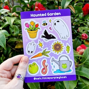 Puede incluir: Una hoja de pegatinas con fondo morado y una variedad de ilustraciones lindas, que incluyen un fantasma, un cuervo, un girasol, una rana, un gusano, una zanahoria, una regadera y una maceta. El texto "Haunted Garden" está en la parte superior de la hoja. El texto "@ash.littlepurpleghost" está en la parte inferior de la hoja.