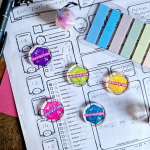 May include: A close-up of a tabletop role-playing game character sheet with several colourful dice with the word "Inspiration" printed on them. The dice are placed on the character sheet next to the Strength, Constitution, Intelligence, Wisdom, and Charisma stats.